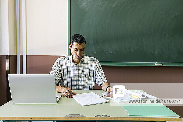 Professor sitting with laptop and checking paper in classroom