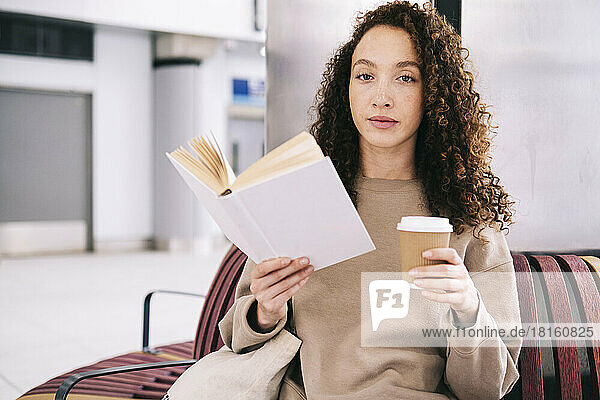 Young woman reading book with disposable cup of coffee