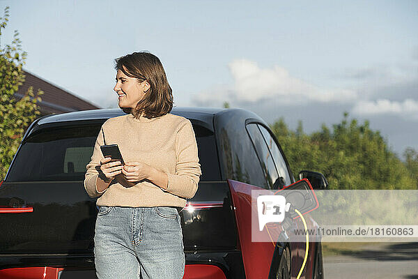 Happy woman with smart phone leaning on car electric at charging station