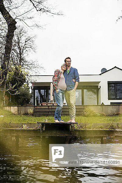 Pregnant woman embracing mature man on jetty in front of house