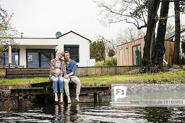 Expectant couple sitting on jetty over lake in front of house