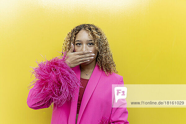 Young woman covering mouth with hand in pink fur blazer against yellow background
