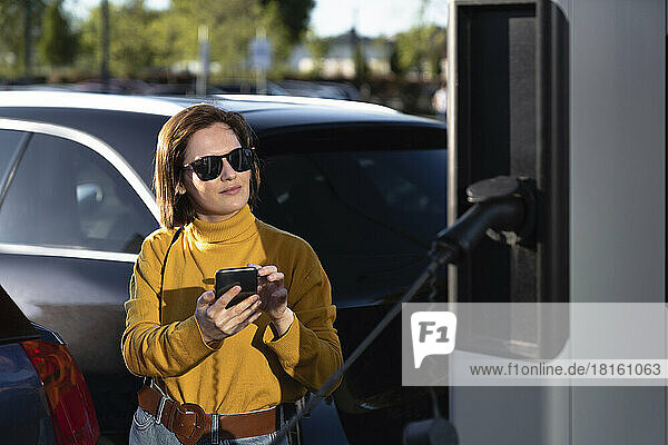 Woman wearing sunglasses standing with mobile phone at vehicle charging station