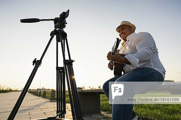 Smiling street musician playing guitar and recording through smart phone on tripod