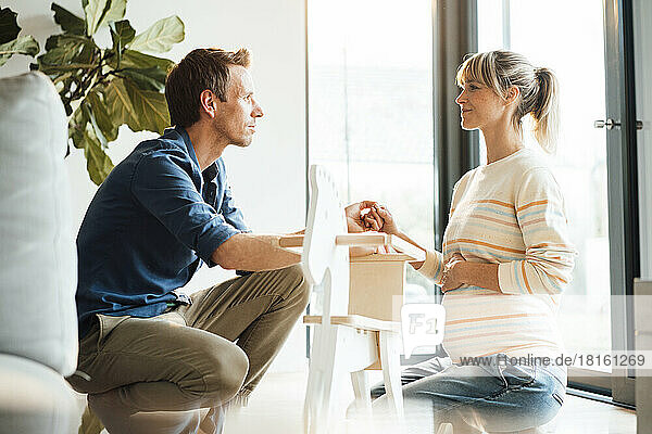 Romantic man holding hand of pregnant woman crouching near rocking horse at home