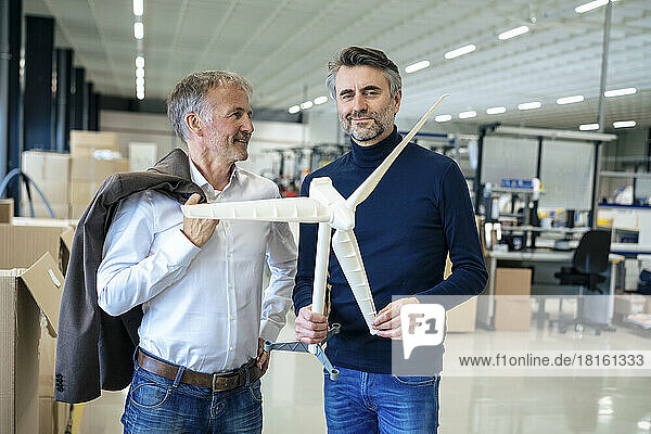 Smiling businessman holding wind turbine model standing by colleague at warehouse