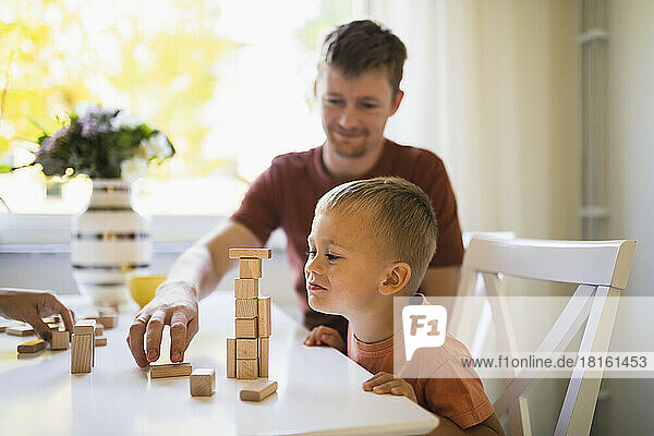 Netter Junge schaut seinem Vater dabei zu  wie er Bauklötze auf dem Esstisch stapelt