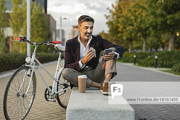 Lächelnder Geschäftsmann benutzt Smartphone und isst mit dem Fahrrad auf der Bank zu Mittag