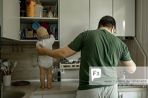 Baby girl standing on kitchen counter with father making coffee