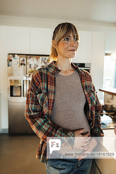 Nachdenkliche schwangere Frau mit den Händen auf dem Bauch steht zu Hause