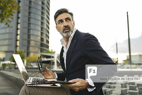 Businessman looking away with laptop sitting on wall