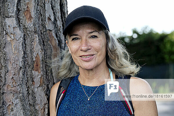 Smiling senior woman wearing cap by tree trunk