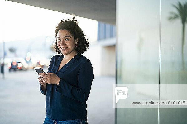 Happy businesswoman with mobile phone standing by glass wall