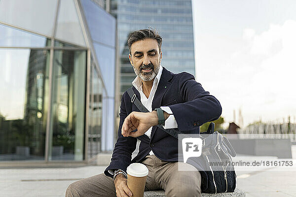 Mature businessman checking time on wristwatch sitting in front of office building