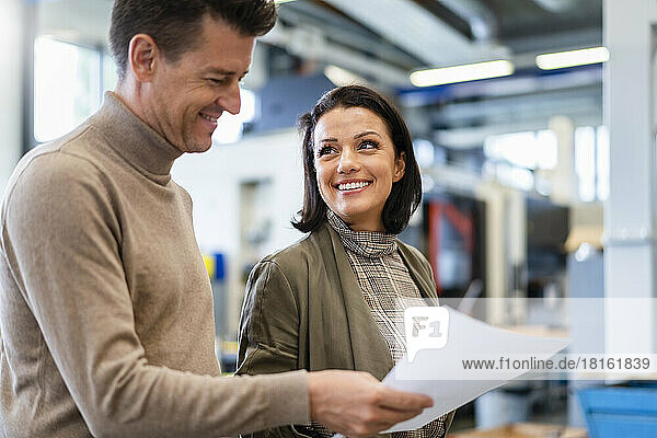 Lächelnde Geschäftsfrau blickt auf Geschäftsmann  der Papier in der Produktionshalle hält