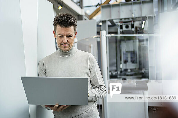 Businessman using laptop leaning on wall in industry