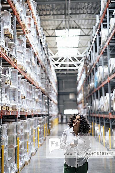Businesswoman holding smart phone standing with hand in pocket at factory