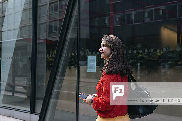 Smiling woman holding mobile phone walking by glass wall
