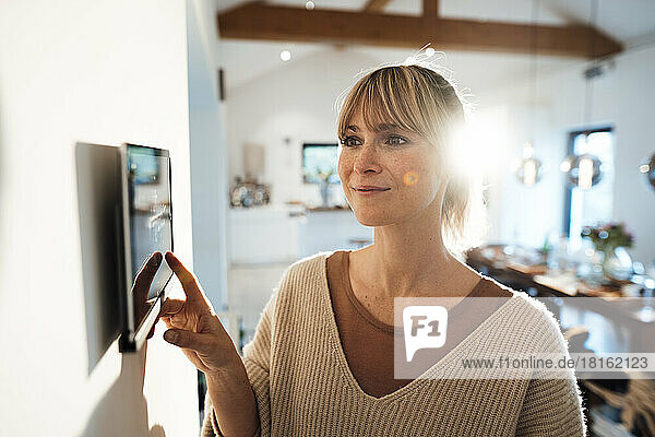 Smiling woman using smart home app through tablet PC mounted on wall at home