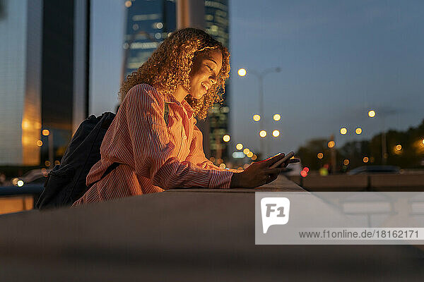 Smiling woman leaning on wall and using mobile phone in city