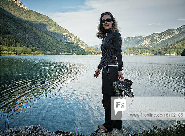 Smiling woman holding boots standing on rock at lake