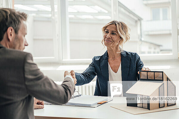 Businesswoman by house model shaking hands with client at office
