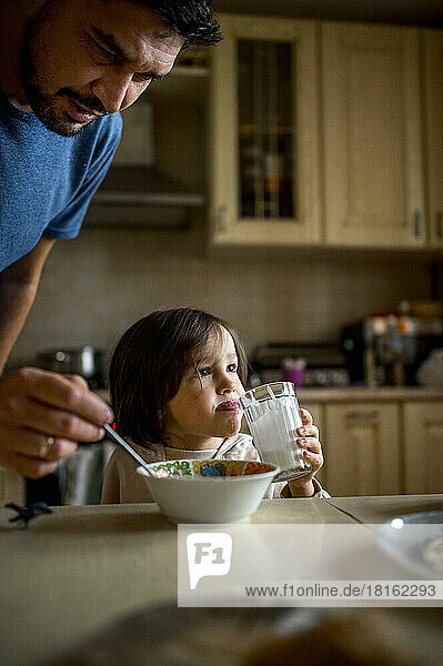 Man feeding son sitting with glass of milk in kitchen at home