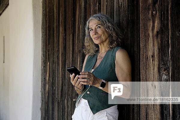 Thoughtful senior woman holding mobile phone leaning on old barn door