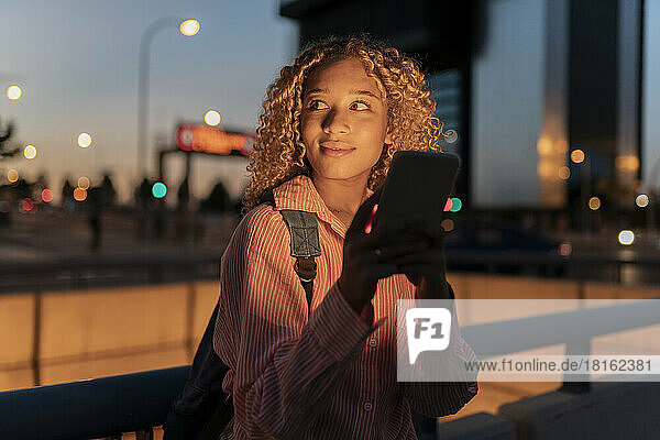 Smiling young woman with smart phone by railing at night