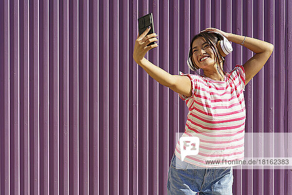 Glückliche junge Frau macht ein Selfie mit ihrem Smartphone vor einer Wellblechwand