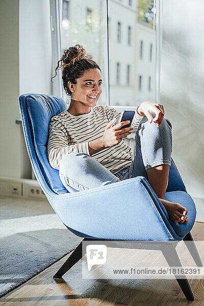 Smiling woman with mobile phone sitting on chair at home