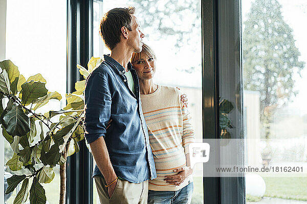 Mature man standing with pregnant woman near glass door at home