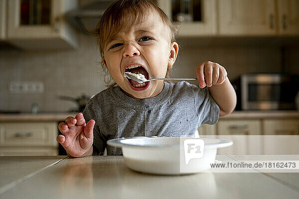 Baby boy eating porridge with spoon in kitchen