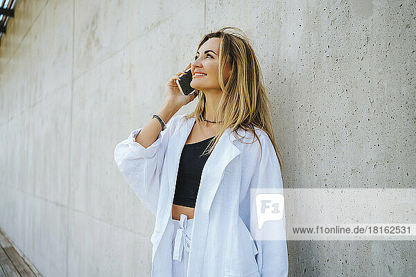 Happy woman talking on smart phone standing in front of wall