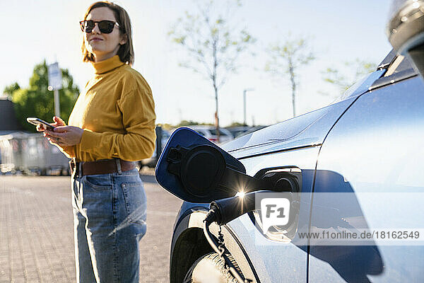Electric car charging at station with woman standing in background on sunny day