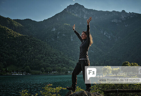 Happy mature woman with arms raised on rock near lake