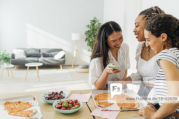 Cheerful roommates having pizza and using smart phone at home