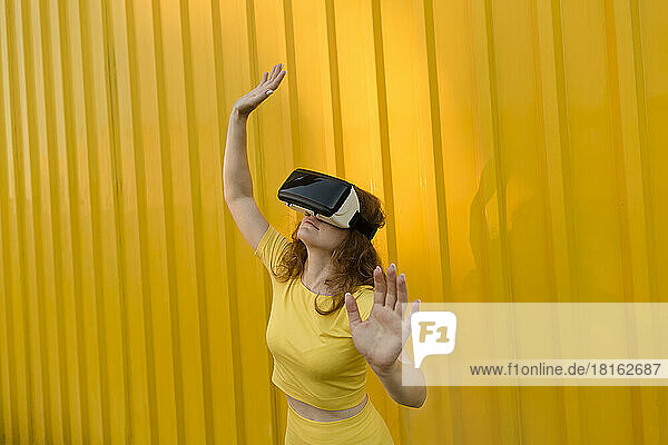 Smiling woman wearing virtual reality simulator gesturing in front of yellow wall