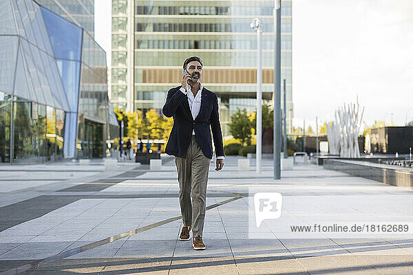 Mature businessman walking and talking on smart phone in front of office building