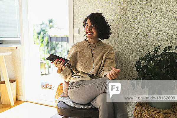 Happy woman holding mobile phone enjoying music with in-ear headphones sitting on chair at home