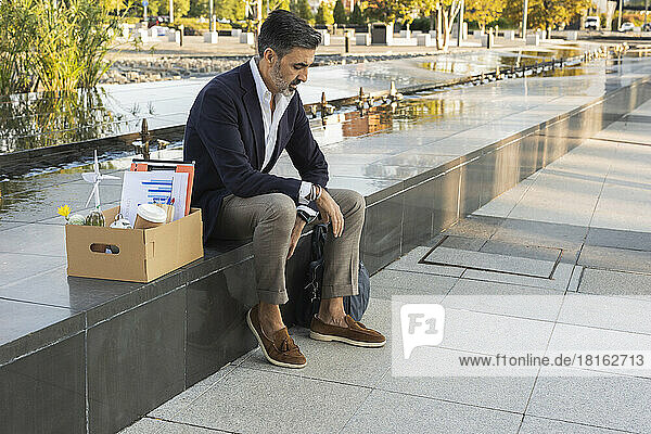 Sad businessman with office supplies in box sitting on wall