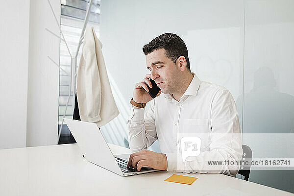 Young businessman talking on smart phone and using laptop at desk in office