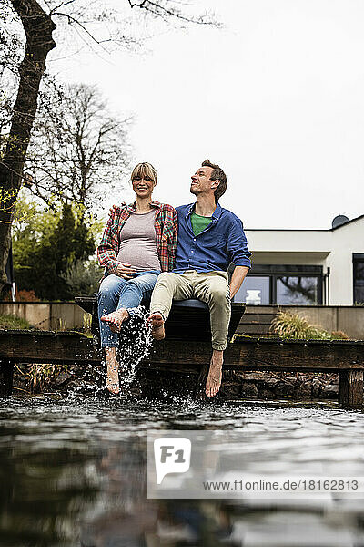 Playful expectant couple splashing water sitting on jetty at lake