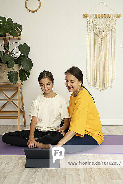 Smiling woman with daughter watching yoga tutorial on tablet PC sitting on exercise mat