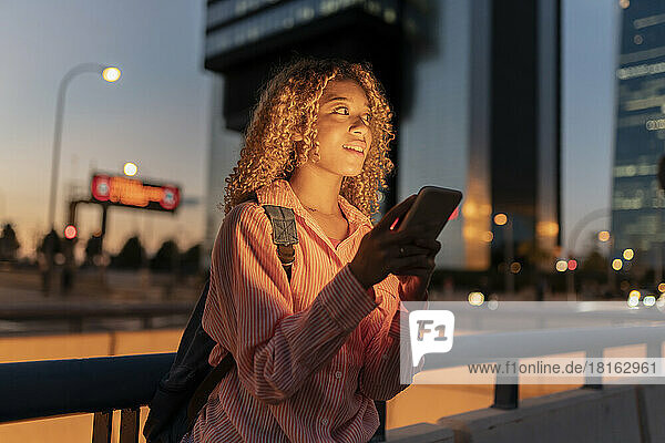 Curly haired woman with mobile phone contemplating at night