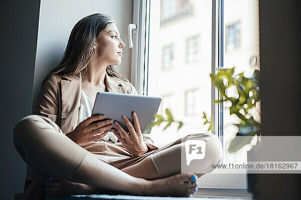 Businesswoman sitting with tablet PC on window sill at home