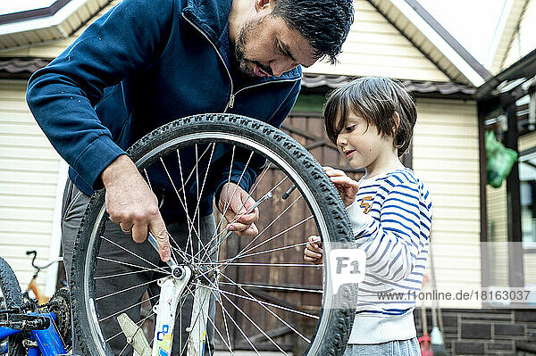Boy looking at father repairing bicycle wheel outside house