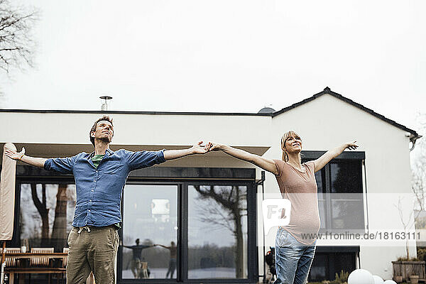 Expectant couple standing with arms outstretched in front of house