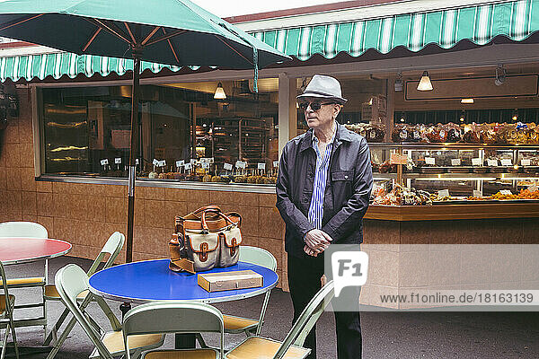 Senior man wearing hat standing by table at sidewalk cafe