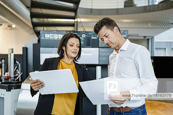 Businesswoman holding tablet PC examining paper standing by colleague in industry